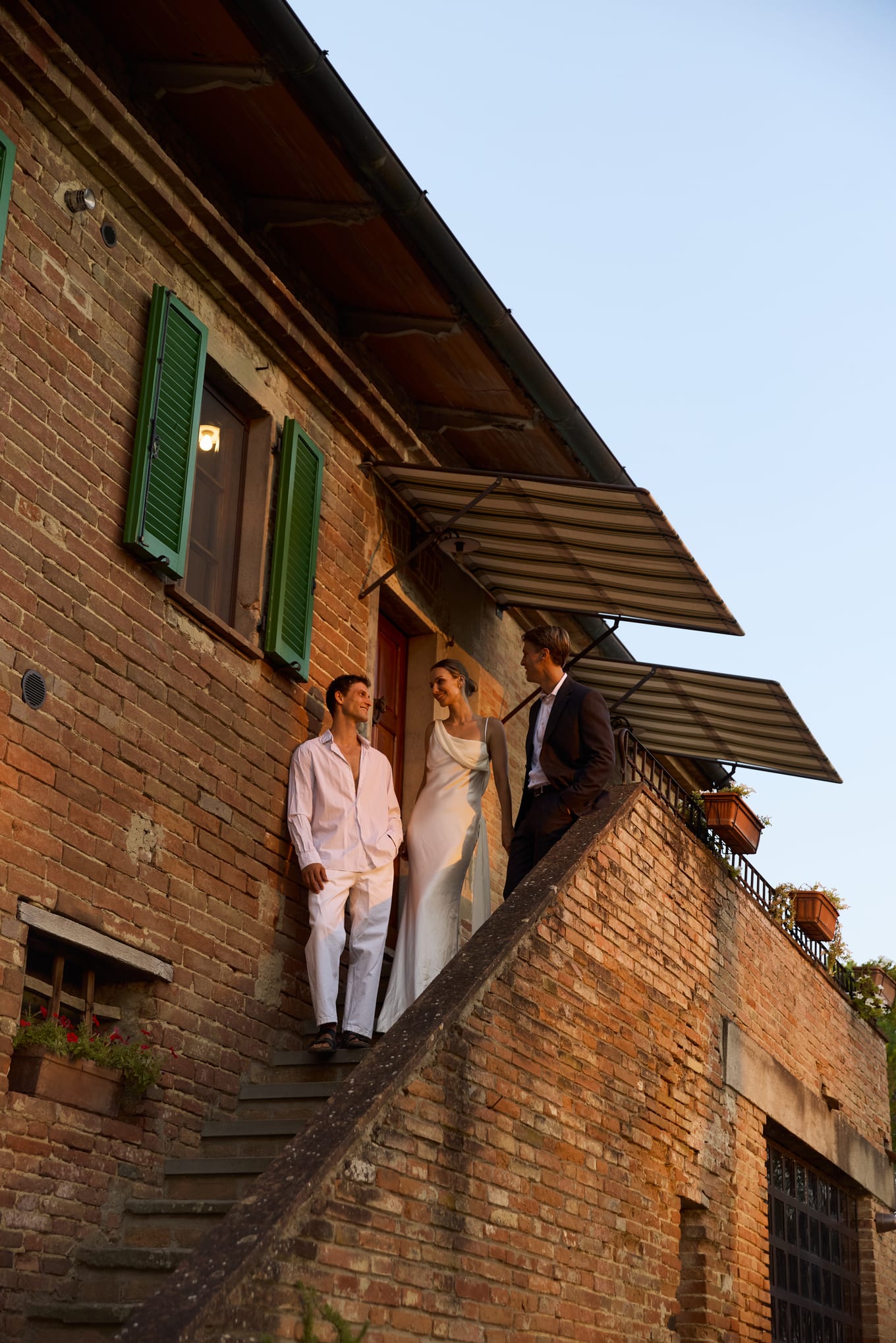 Two men and a woman in a white gown pause on the outdoor stone stairs of a brick farmhouse
