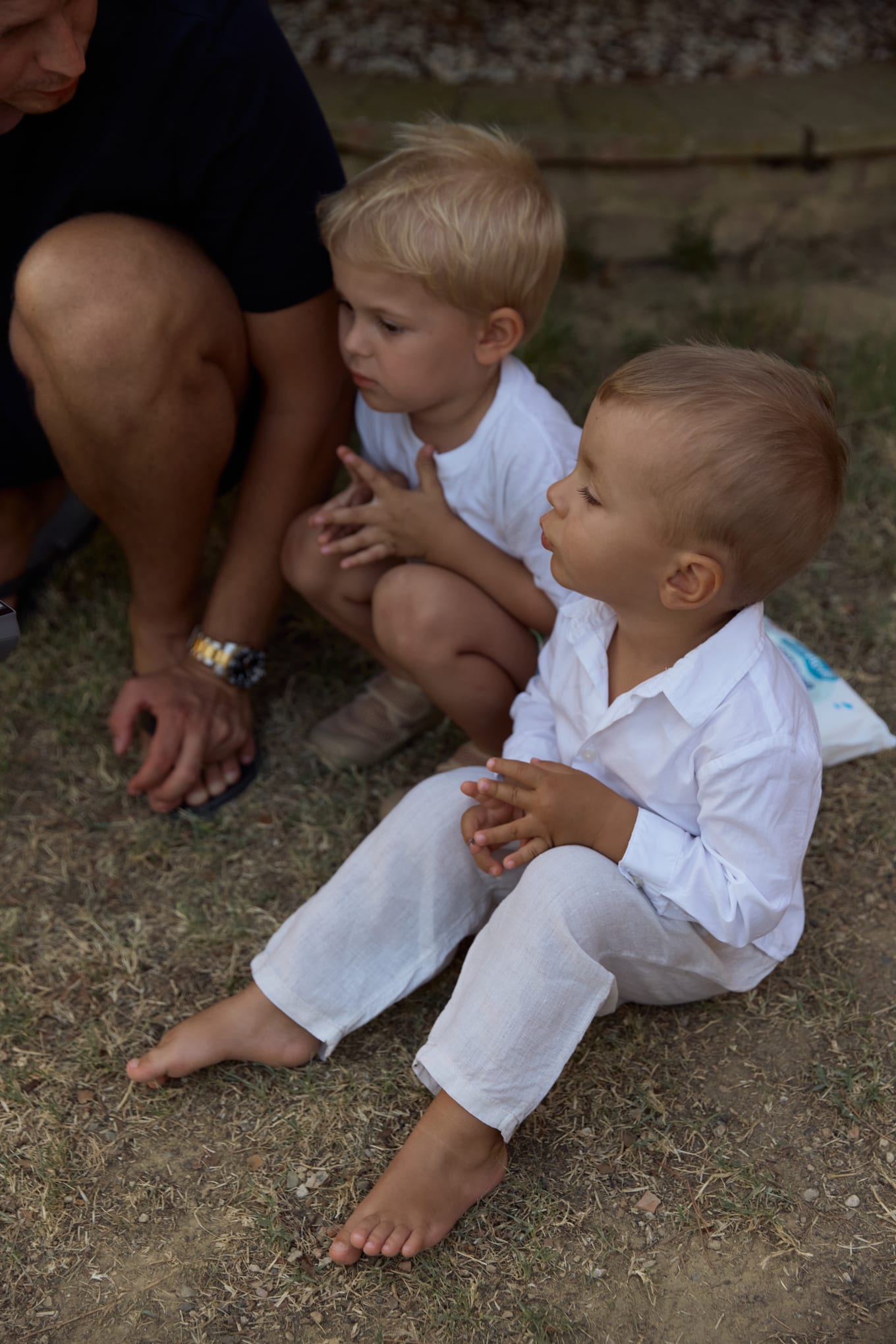 Two small blond boys in white clothes sit barefoot on dry grass beside a crouching man