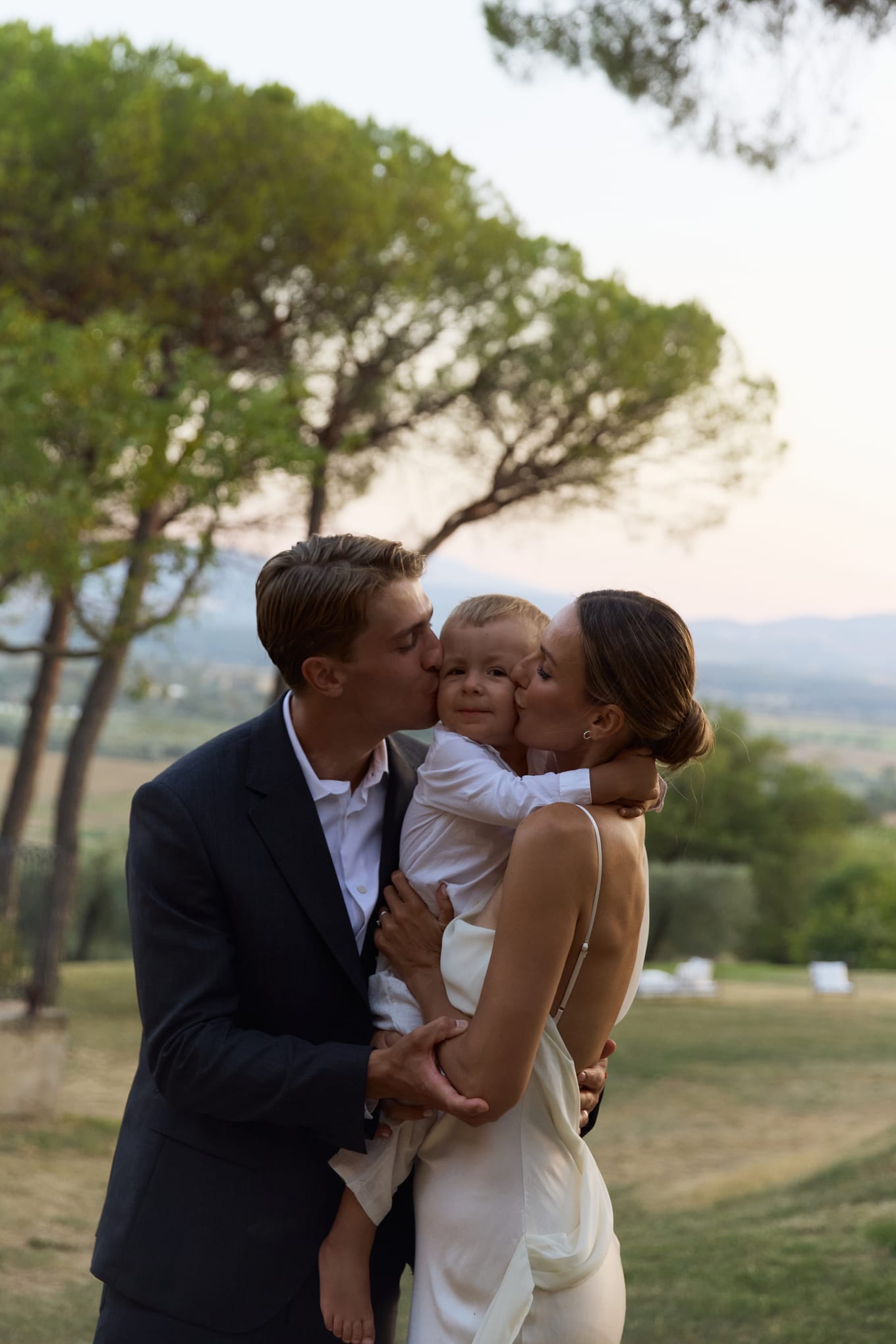 Man in a dark suit and woman in a white slip dress both kiss the cheeks of a toddler held between them