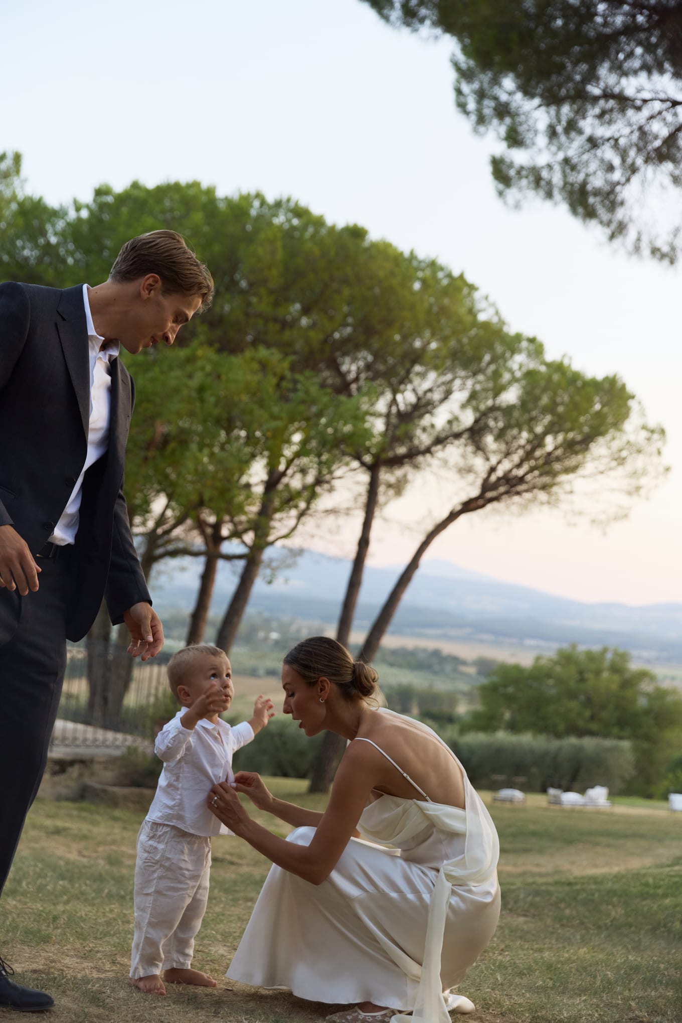 Barefoot toddler in white stands on the grass as a woman crouches to hold his hands and a man looks down