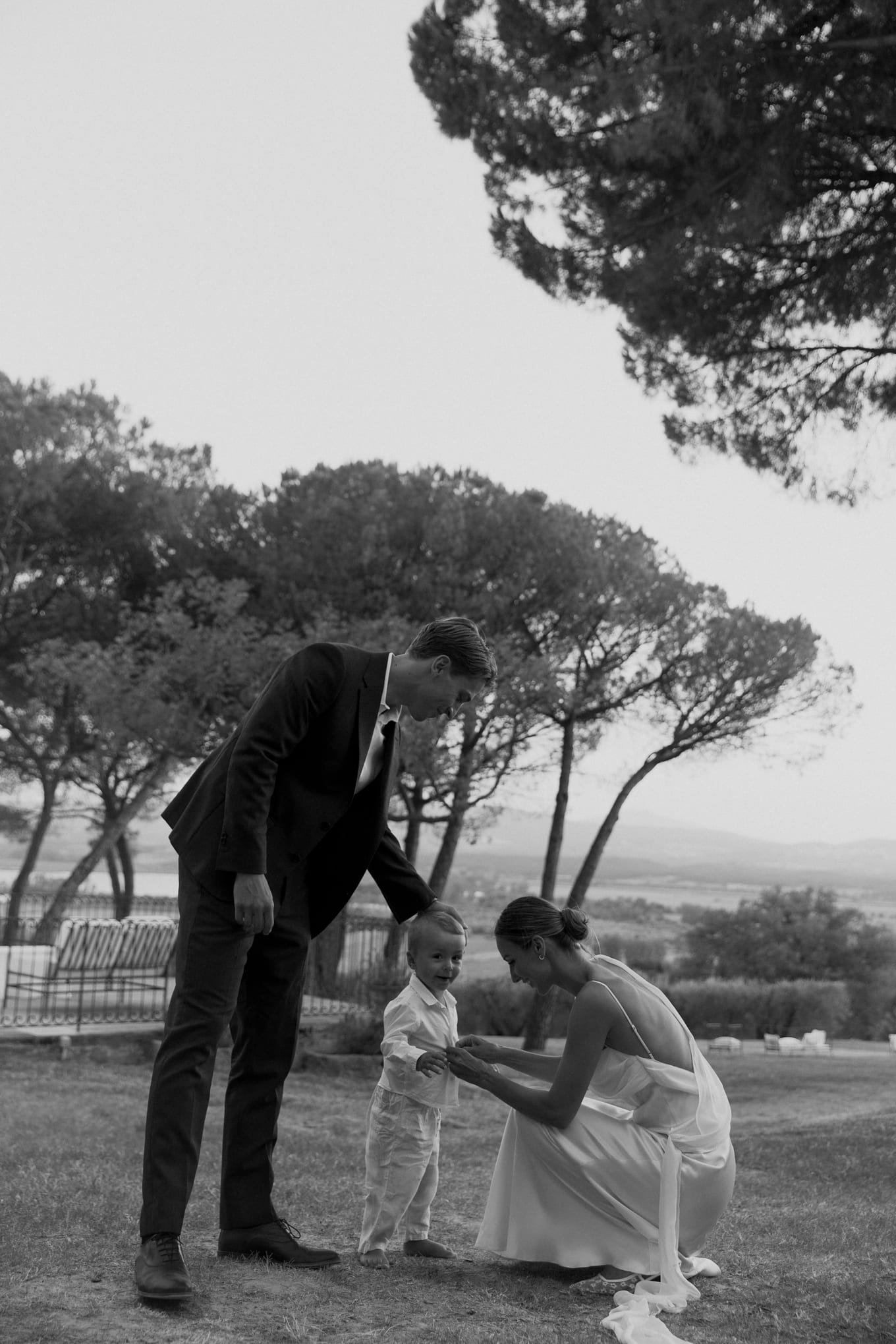 Under tall pines in black and white, a woman kneels to hold a small boy's hands while a man leans over them