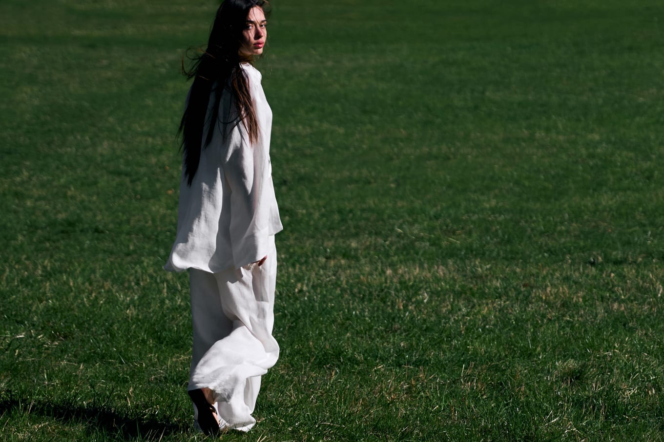 Long-haired woman in a loose white linen outfit walks across a sunny lawn, glancing back over her shoulder