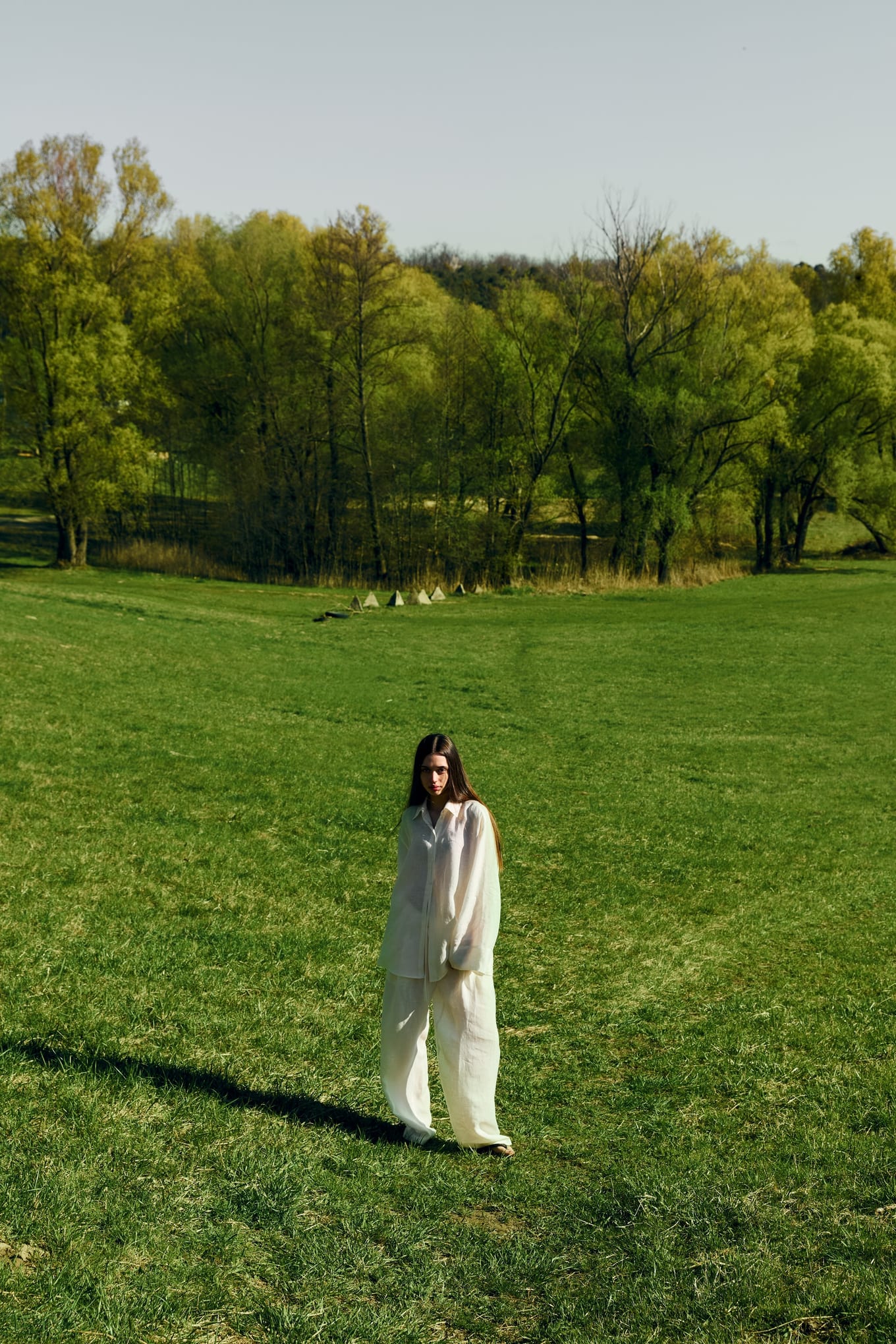 Woman in white linen shirt and trousers stands alone in a wide green meadow with trees behind her