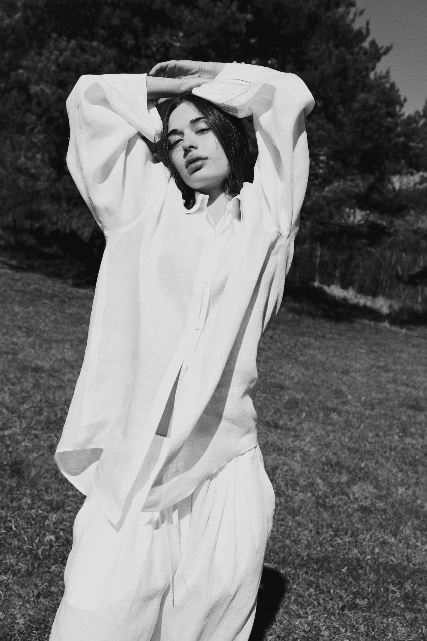 In black and white, a woman in loose white linen rests her hands on her head, eyes half closed, on grass
