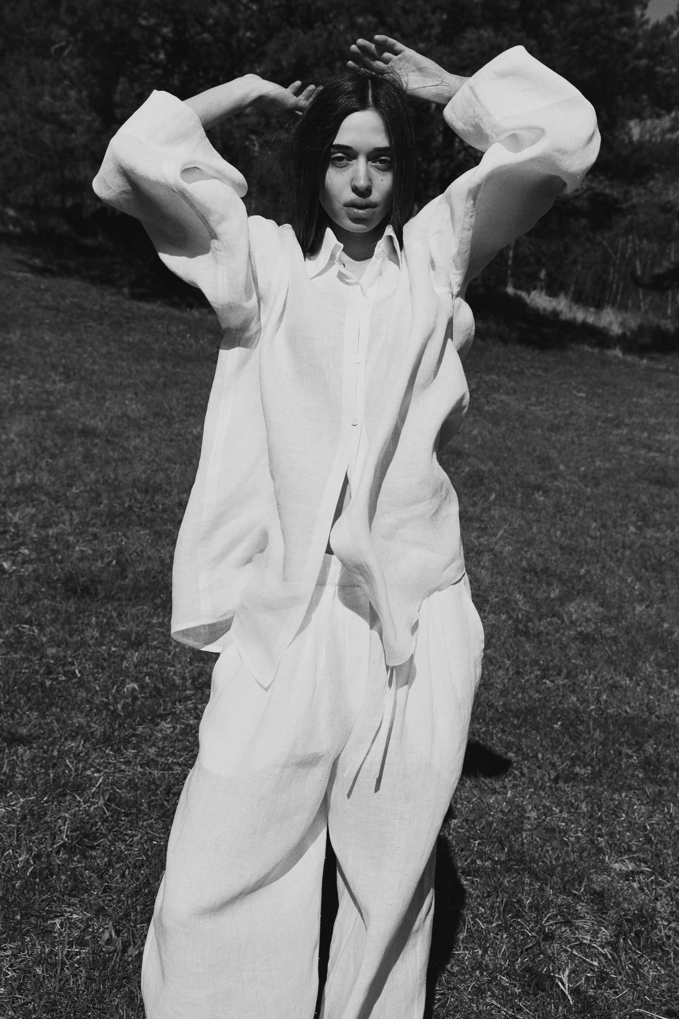 Woman in a loose white linen shirt and trousers stands on grass with both hands raised to her head