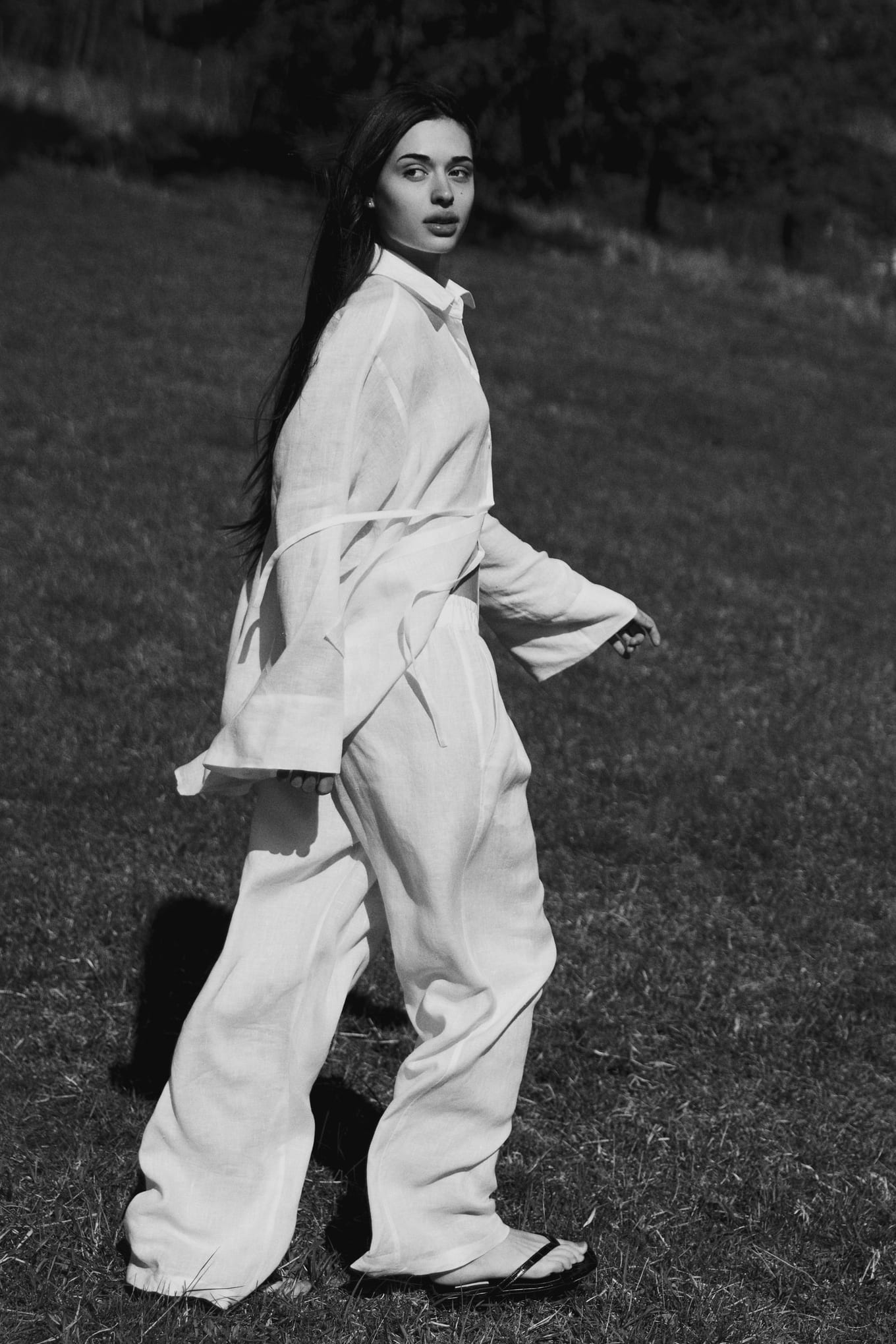 Walking across a grassy slope, a woman in a white shirt and wide trousers glances over her shoulder