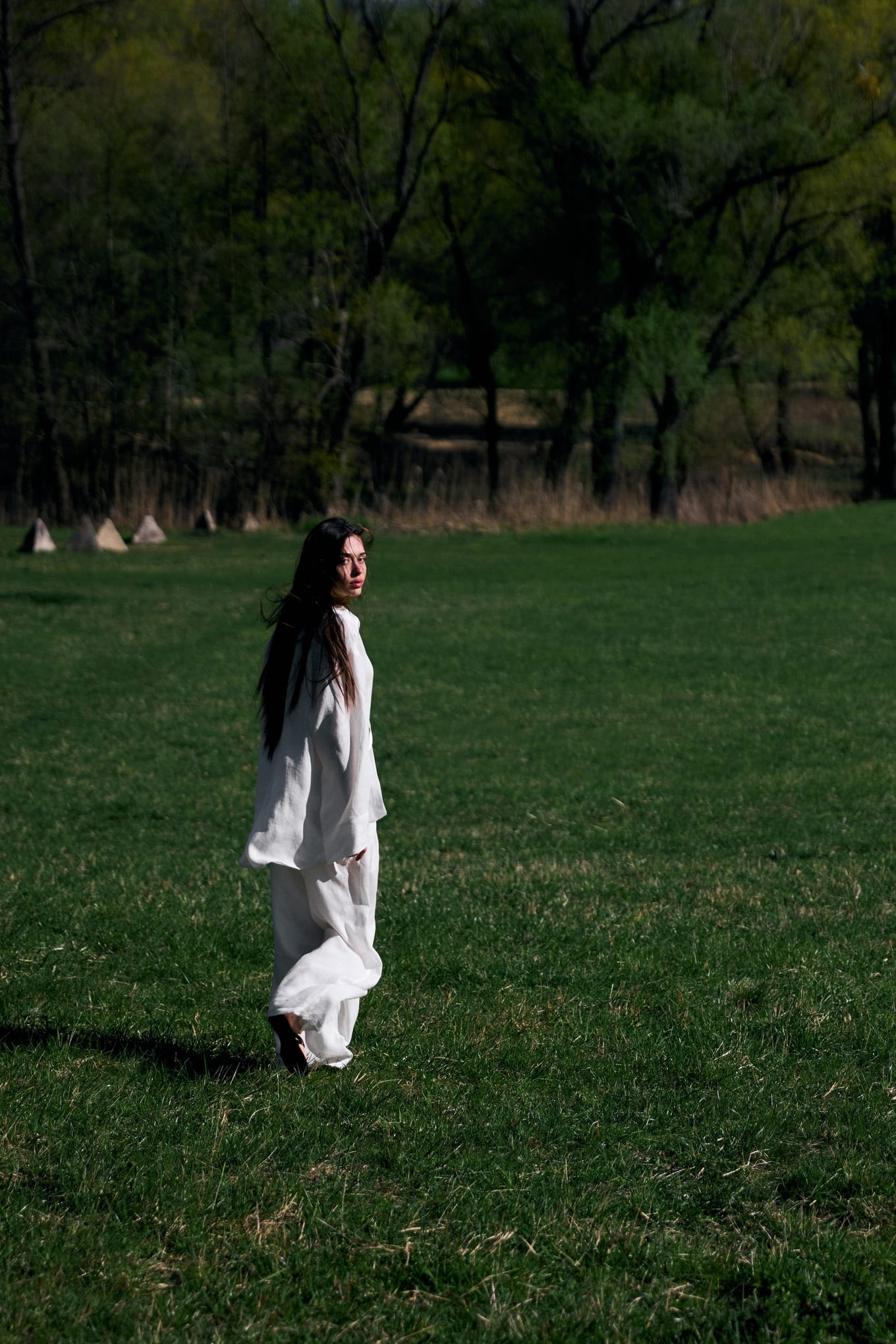 Seen from a distance, a woman in white walks across a green field toward a line of trees