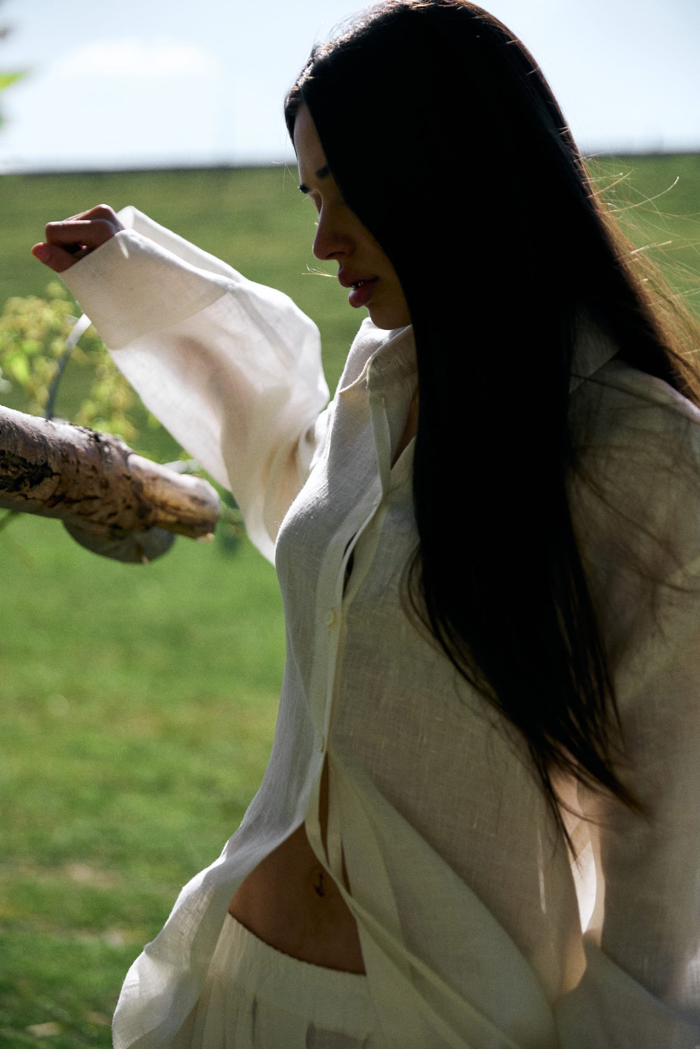 In profile, a woman with long dark hair and an open linen shirt reaches toward a branch in sunlight