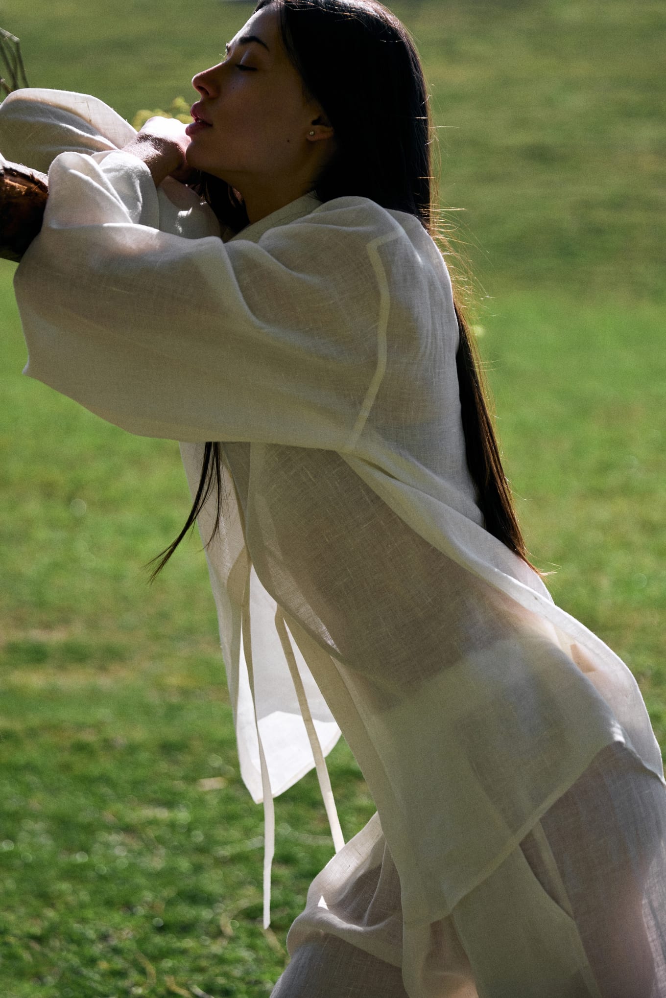 Dark-haired woman in a sheer white linen shirt leaning forward with eyes closed on a sunlit lawn