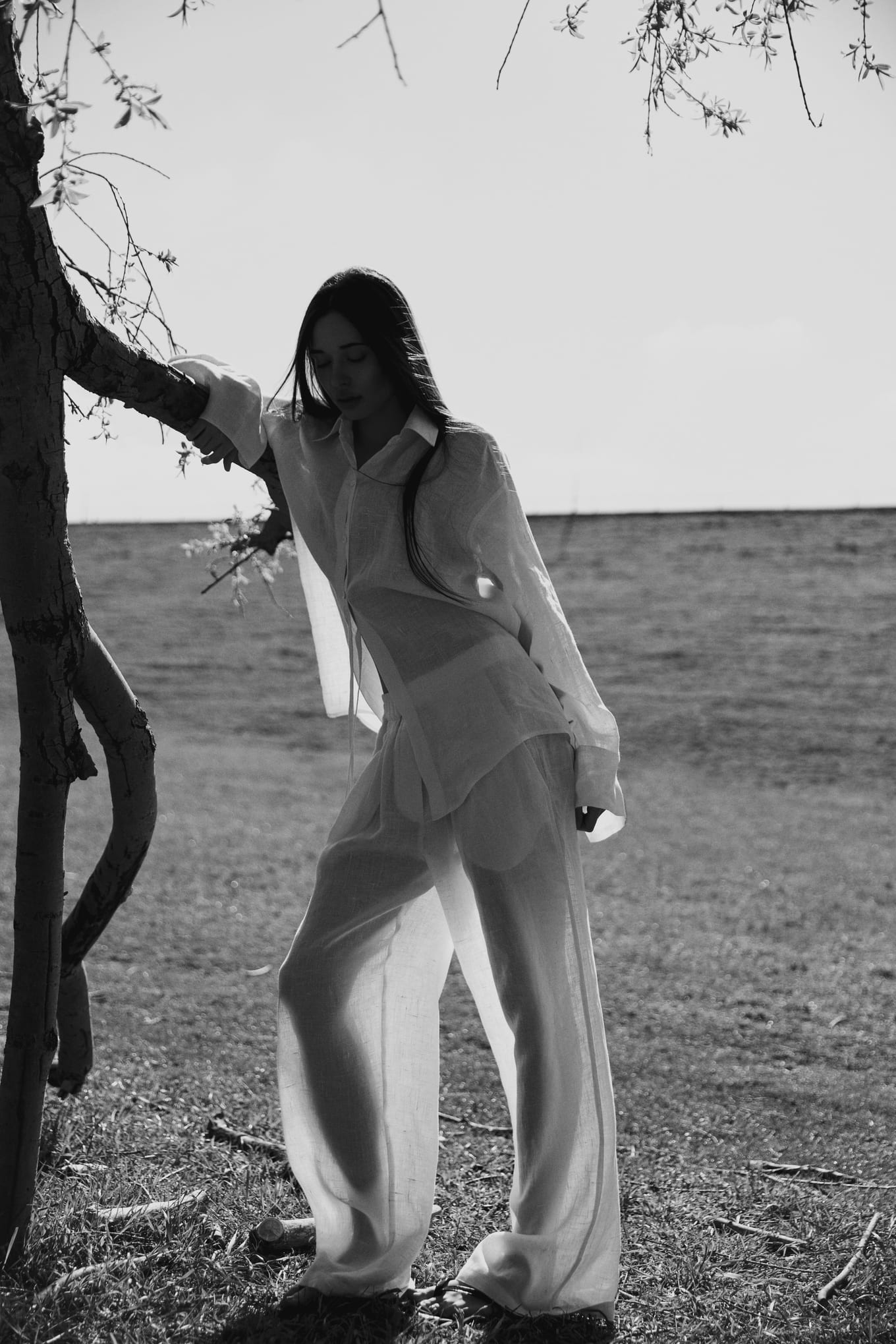 Black and white shot of a woman in sheer linen leaning on a slender tree trunk in an open field