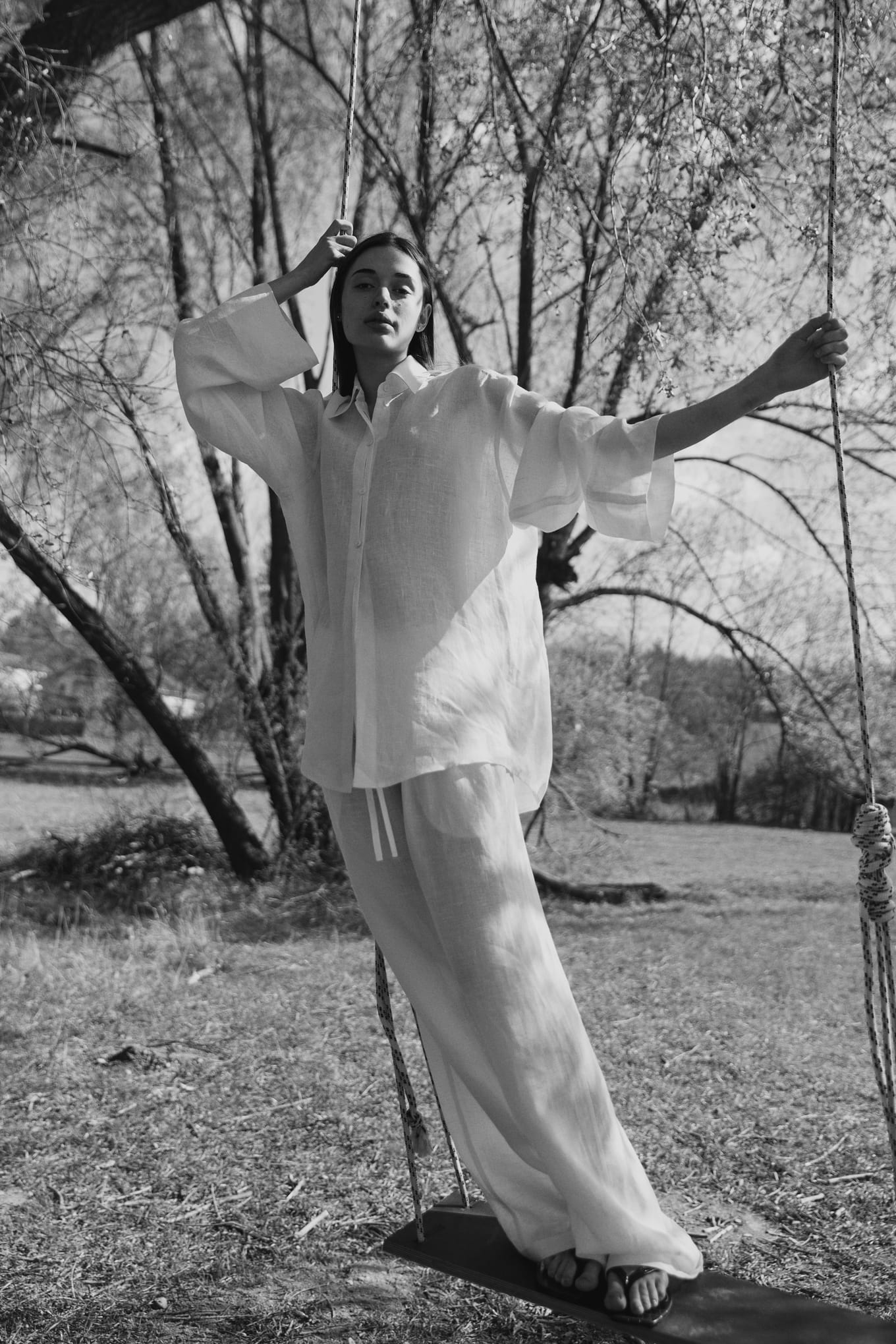 Woman in loose white linen standing on a rope swing among bare trees, one hand raised to her head