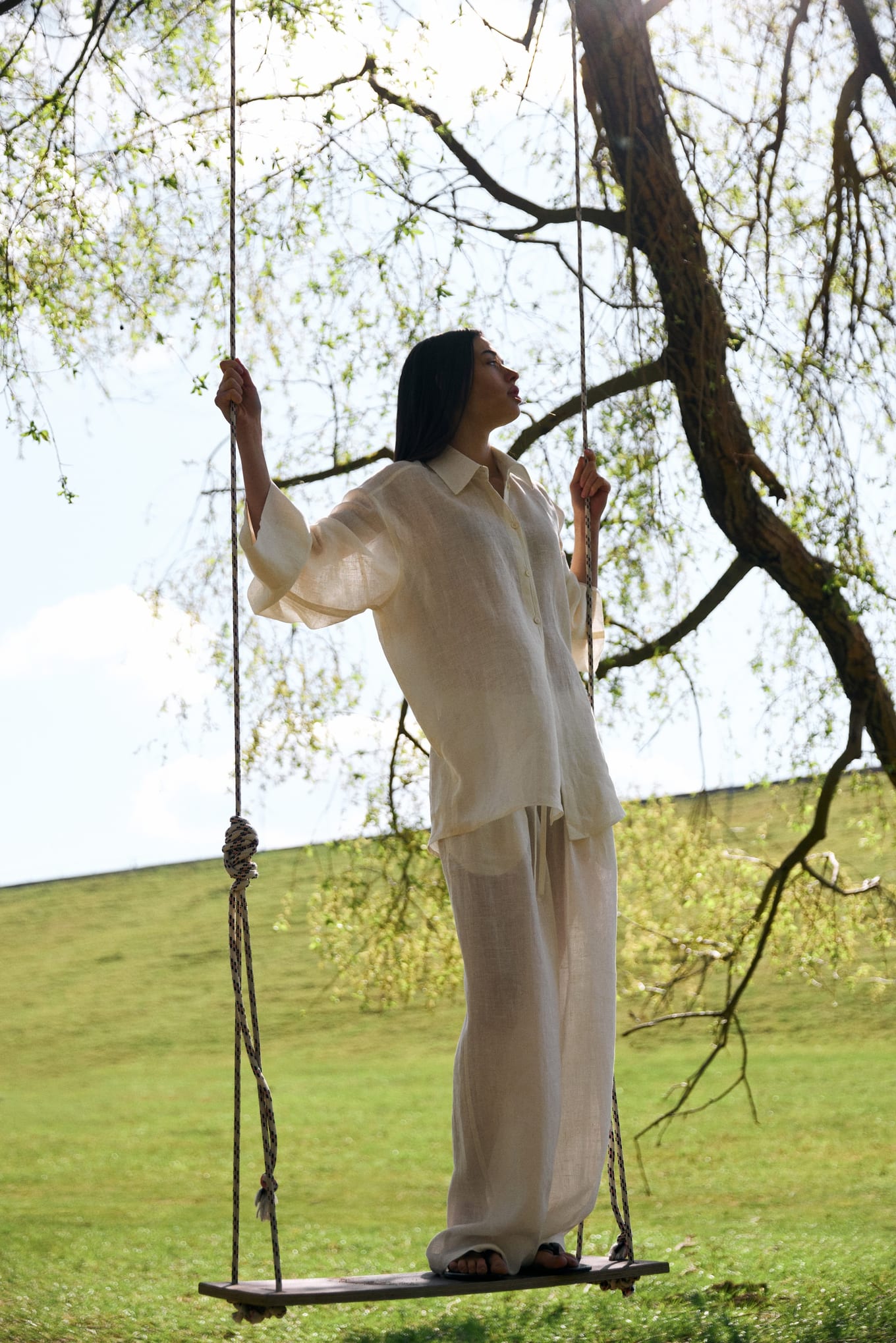 Standing on a wooden swing beneath a leafy tree, a woman in cream linen holds both ropes and looks up