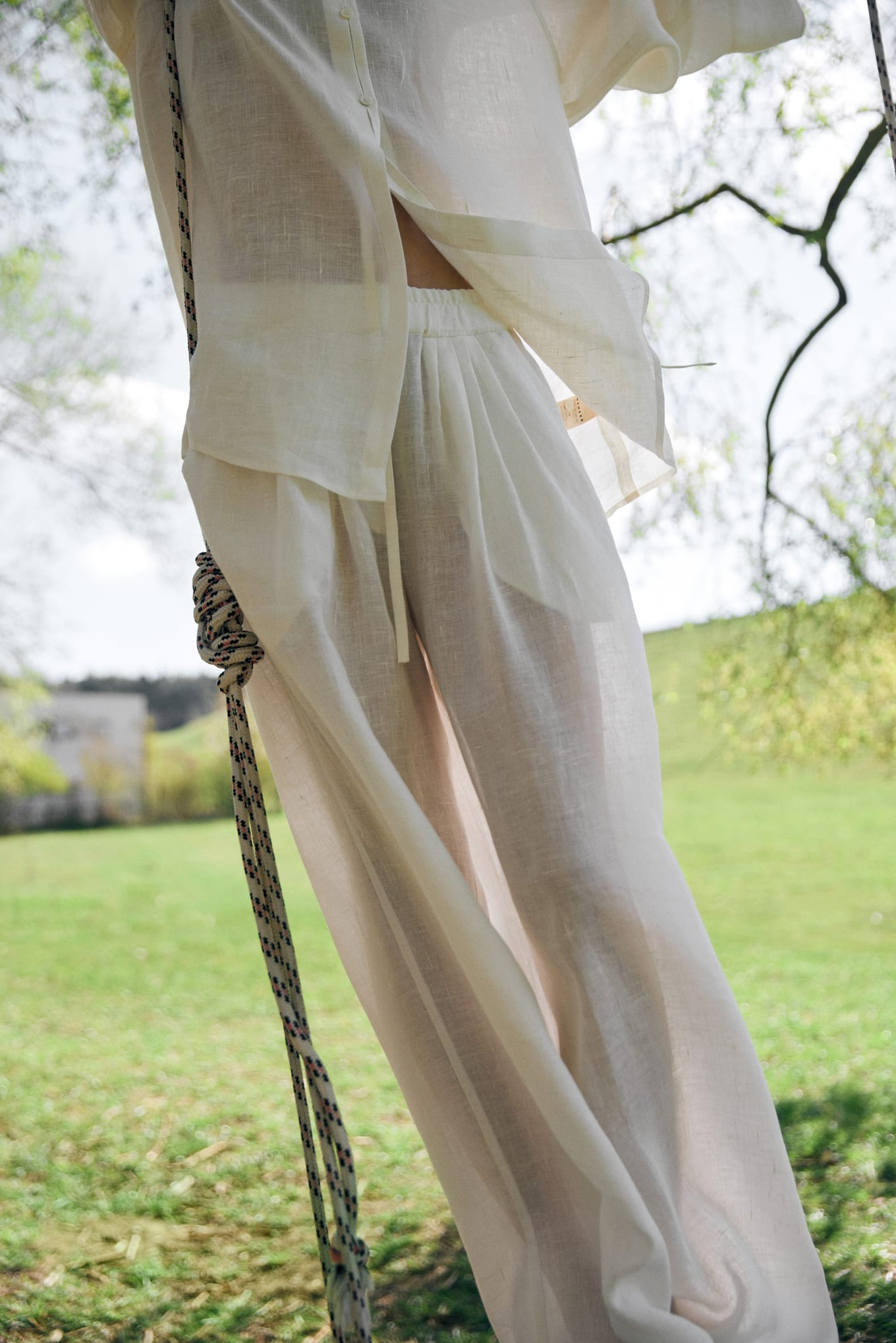 Cropped view of wide sheer linen trousers and a loose shirt beside a knotted swing rope on grass