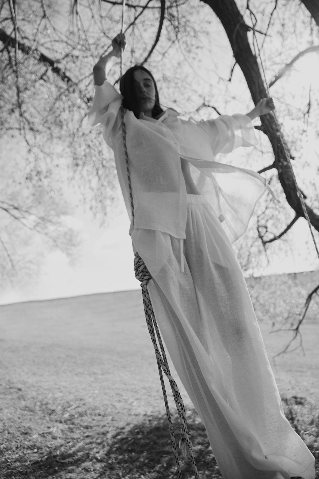 Woman in sheer white shirt and wide trousers holds the ropes of a tree swing, hair blowing across her face