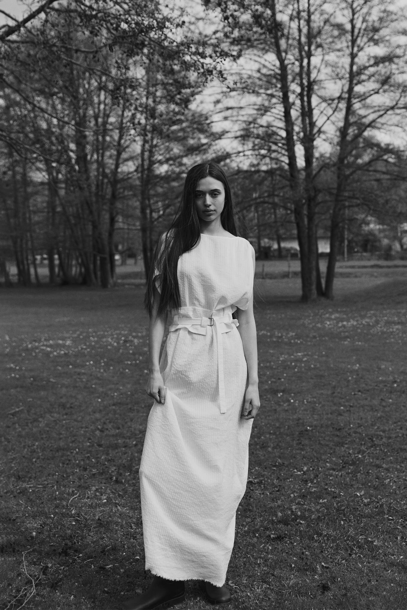 Standing on grass beneath tall trees, a woman in a long white belted dress looks toward the camera
