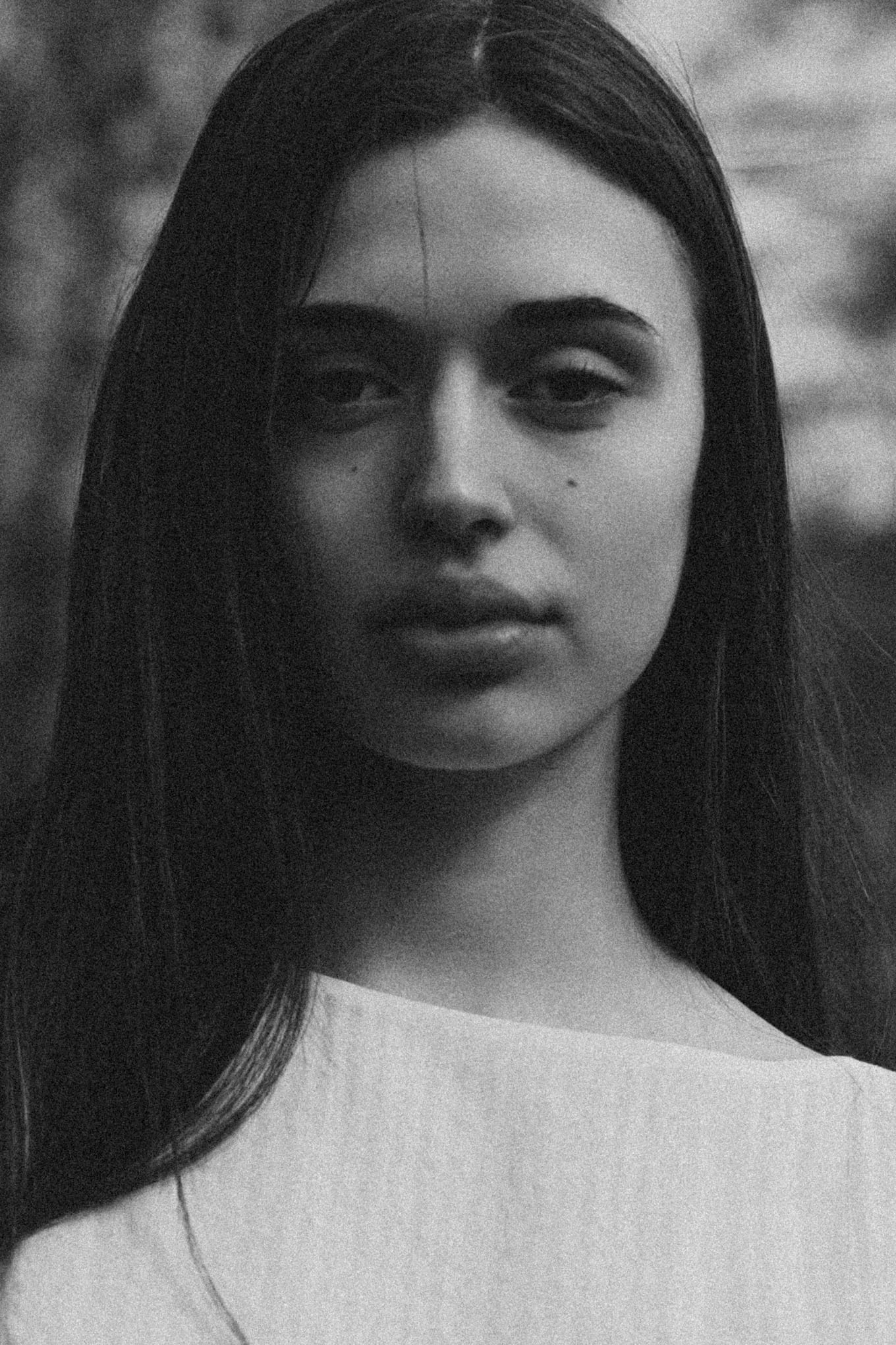 Close black-and-white portrait of a young woman with long dark hair in a pale top, trees blurred behind