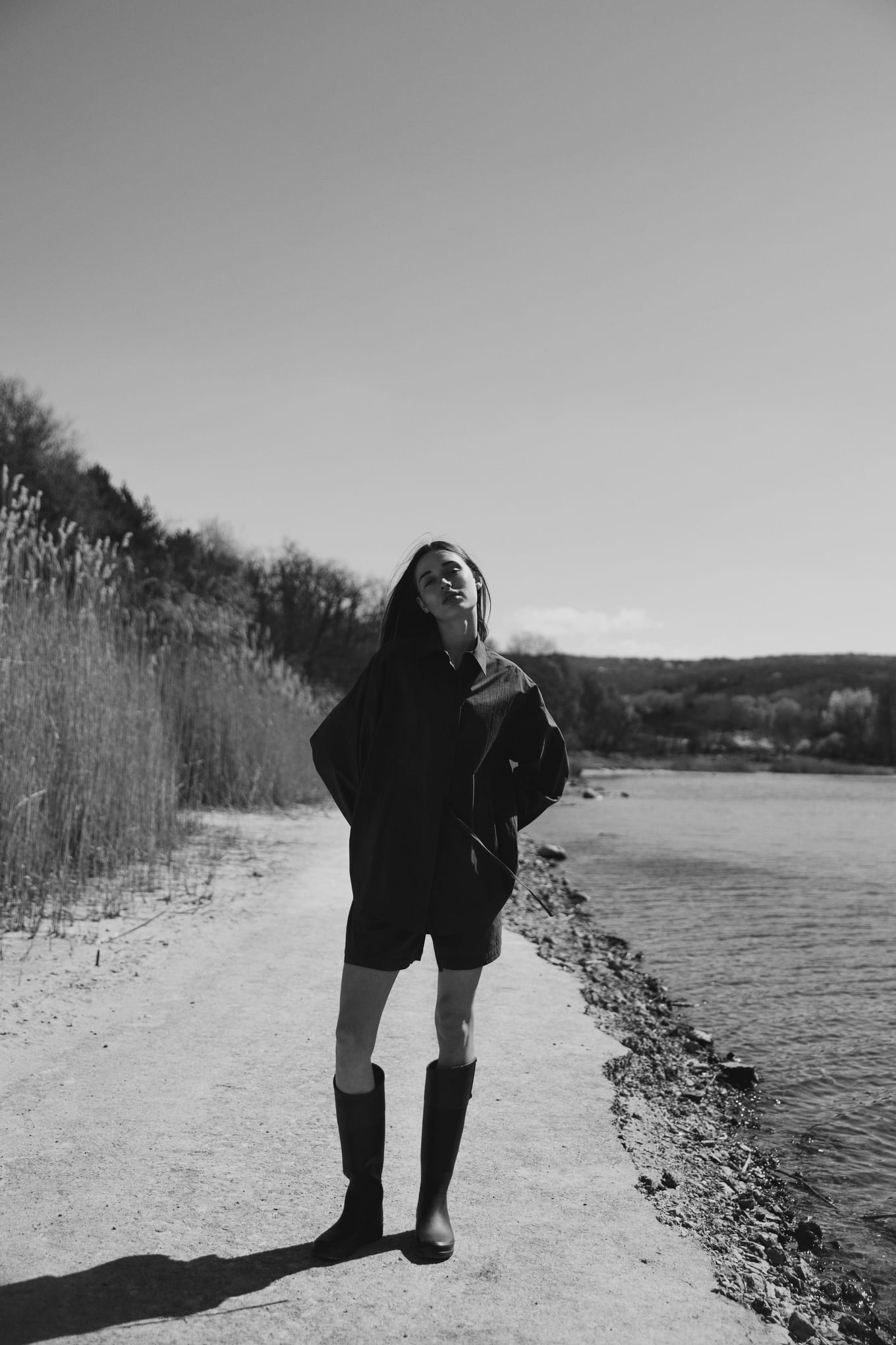 On a sandy path between tall reeds and a lake, a woman in a dark shirt and tall boots tilts her head back