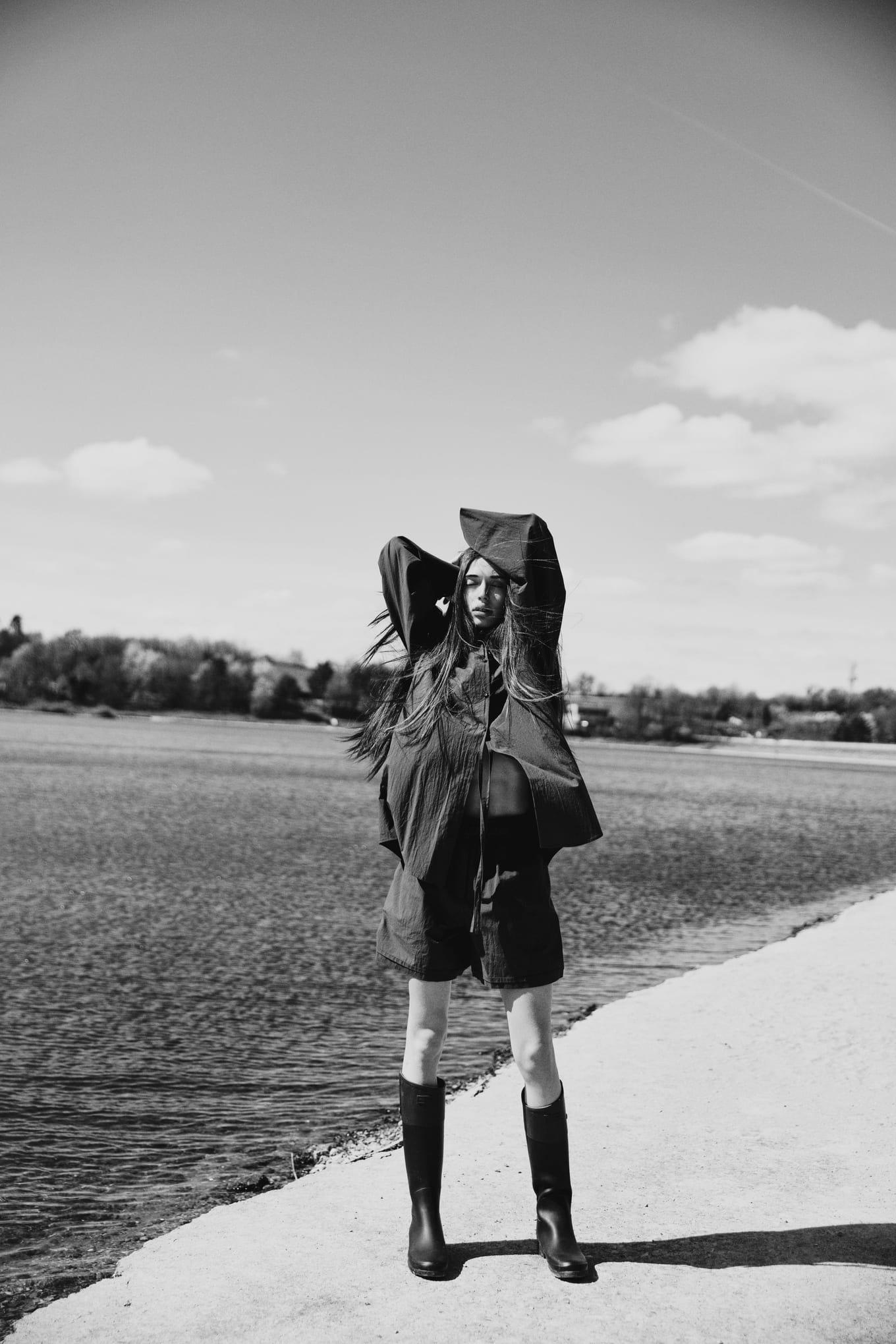 Hair blowing in the wind, a woman lifts the collar of a dark jacket over her head on a lakeside walkway