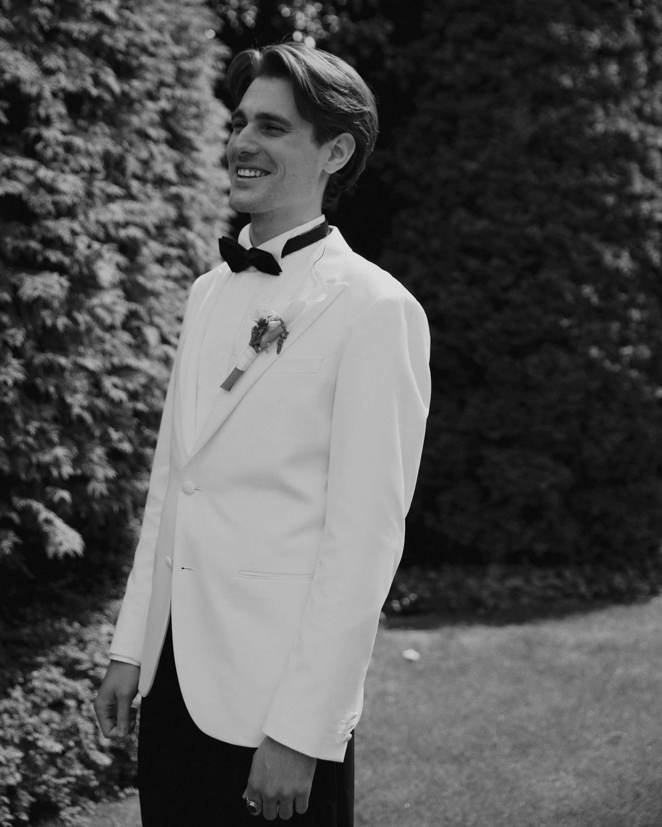 Groom in a white dinner jacket beside a hedge