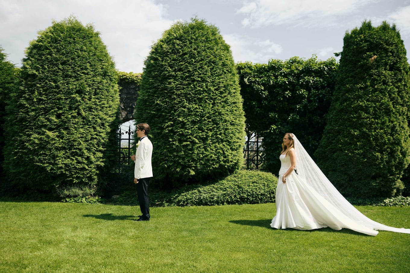 First look between clipped hedges, the bride's train across the grass