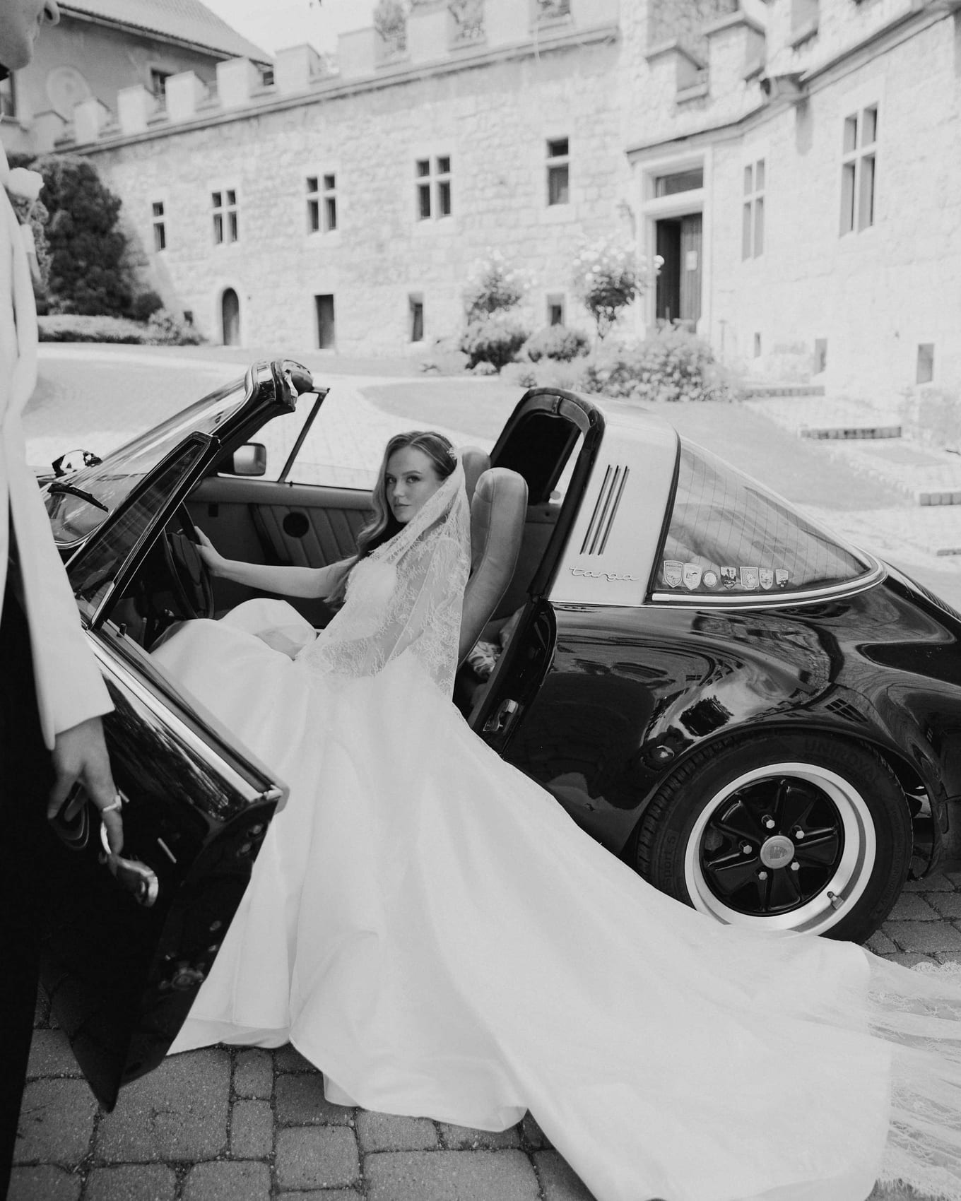 Bride seated in the open car with her skirt across the cobbles